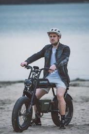 A person wearing a helmet sits on an electric bike on a sandy beach with a large body of water in the background. The bike has thick tires and the brand name Surpanda is visible on its frame. The person is dressed in a casual jacket, shorts, and sneakers.
