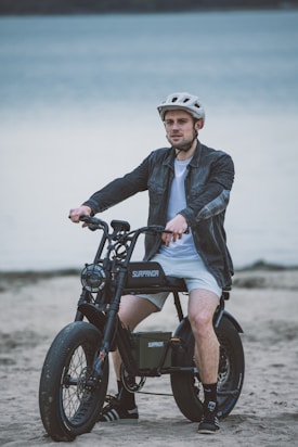 A person wearing a helmet sits on an electric bike on a sandy beach with a large body of water in the background. The bike has thick tires and the brand name Surpanda is visible on its frame. The person is dressed in a casual jacket, shorts, and sneakers.