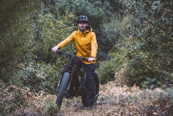 A person wearing a yellow jacket and black helmet is standing with a mountain bike on a narrow, winding path surrounded by dense green foliage. The scene suggests an outdoor, natural setting, possibly a forest or wooded area. The person appears to be prepared for an adventure, shown by their gear and the rugged terrain.