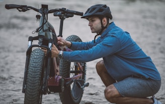 A person wearing a helmet is kneeling on one knee in an outdoor setting. They are adjusting or examining a large, off-road bicycle with thick tires. The individual is dressed in a blue denim shirt and shorts, suggesting a casual activity. The surface appears sandy, indicating a beach or similar environment.
