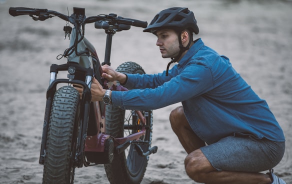 A person wearing a helmet is kneeling on one knee in an outdoor setting. They are adjusting or examining a large, off-road bicycle with thick tires. The individual is dressed in a blue denim shirt and shorts, suggesting a casual activity. The surface appears sandy, indicating a beach or similar environment.