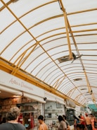 A bustling market scene with several stalls selling arts, decorations, and various goods. The market is sheltered by a large, curved canopy with a yellow framework. Numerous people are browsing the stalls, creating a lively atmosphere.