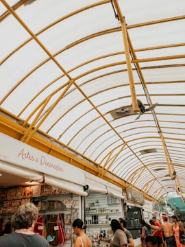 A bustling market scene with several stalls selling arts, decorations, and various goods. The market is sheltered by a large, curved canopy with a yellow framework. Numerous people are browsing the stalls, creating a lively atmosphere.