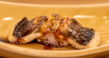 A piece of sushi with grilled fish on top, placed on a light brown plate. The fish is glazed, giving it a shiny appearance. Underneath the fish is a mound of rice. The overall presentation is elegant and appetizing.