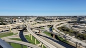 A complex network of elevated highways and overpasses forms an intricate pattern amidst a sprawling urban area. Multiple lanes of traffic are visible, with vehicles traveling in different directions. The cityscape extends into the distance with numerous buildings and infrastructure, and there's a clear blue sky above.