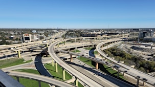 A complex network of elevated highways and overpasses forms an intricate pattern amidst a sprawling urban area. Multiple lanes of traffic are visible, with vehicles traveling in different directions. The cityscape extends into the distance with numerous buildings and infrastructure, and there's a clear blue sky above.