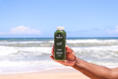 A hand is holding a bottle of green juice against the backdrop of a beach with an expansive view of the ocean and clear blue sky. The label on the bottle is visible with text detailing the contents.