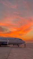 A commercial airplane is parked on the tarmac during sunrise or sunset, with a striking sky filled with vibrant orange and pink hues. The aircraft has visible text in a foreign language and an airline logo. A person in reflective gear stands near the nose of the plane.