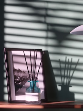 A blue glass vase with several reed diffuser sticks sits on a wooden surface. Behind it, there is a framed black and white photograph. Horizontal shadows from blinds create a pattern on the wall and objects.