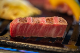 A thick, juicy slice of marbled meat is being grilled to brown perfection. The meat has a distinct pink and white marbling, showcasing its freshness and quality. It rests on a grill with other blurred food items in the background, suggesting a cooking process.