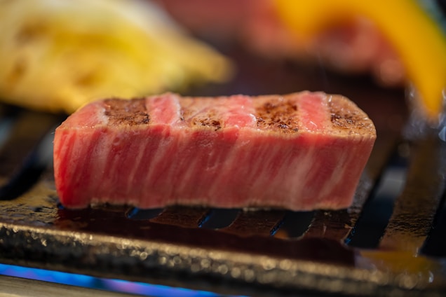 A thick, juicy slice of marbled meat is being grilled to brown perfection. The meat has a distinct pink and white marbling, showcasing its freshness and quality. It rests on a grill with other blurred food items in the background, suggesting a cooking process.