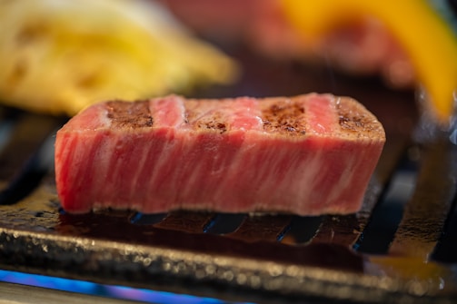 A thick, juicy slice of marbled meat is being grilled to brown perfection. The meat has a distinct pink and white marbling, showcasing its freshness and quality. It rests on a grill with other blurred food items in the background, suggesting a cooking process.
