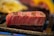 A thick, juicy slice of marbled meat is being grilled to brown perfection. The meat has a distinct pink and white marbling, showcasing its freshness and quality. It rests on a grill with other blurred food items in the background, suggesting a cooking process.