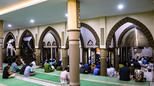 The image depicts a group of people gathered inside a mosque. The architecture features large, ornate arches and columns with intricate details. People are seated on green carpets, some appearing to be in prayer or meditation. The atmosphere is peaceful and reverent.