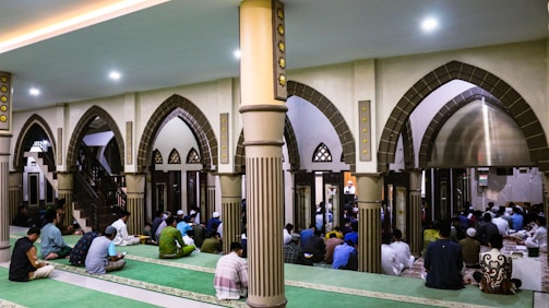 The image depicts a group of people gathered inside a mosque. The architecture features large, ornate arches and columns with intricate details. People are seated on green carpets, some appearing to be in prayer or meditation. The atmosphere is peaceful and reverent.