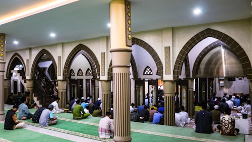 The image depicts a group of people gathered inside a mosque. The architecture features large, ornate arches and columns with intricate details. People are seated on green carpets, some appearing to be in prayer or meditation. The atmosphere is peaceful and reverent.