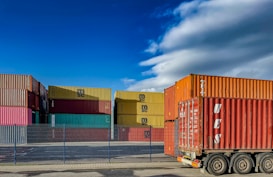 A collection of stacked shipping containers arranged in an industrial area. There are different colors of containers, including red, yellow, pink, and teal. A large orange shipping container is loaded onto a trailer on the right side of the image. The scene is highlighted by a bright blue sky with some clouds.