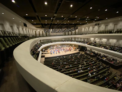 A large concert hall interior with a curved balcony overlooking a stage where an orchestra is performing. The seating area is filled with an audience, and the ambiance is enhanced by a series of overhead lights on the ceiling.