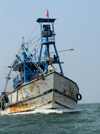 A large fishing boat with a rusty hull navigating through the water. The boat is painted primarily in blue and white, and features a tall structure with people standing on it. A red flag flies at the top of the structure, and several ropes and equipment are visible.