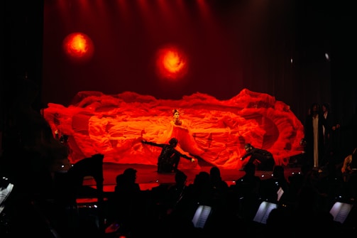 An elaborate stage performance featuring a dramatic red backdrop with fluid, fiery textures resembling flames. Center stage is a performer in an elaborate costume surrounded by other performers in dynamic poses. The lighting enhances the intensity with warm, fiery hues.