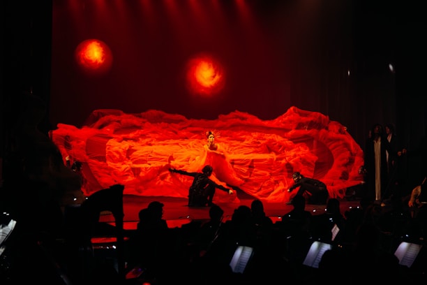 An elaborate stage performance featuring a dramatic red backdrop with fluid, fiery textures resembling flames. Center stage is a performer in an elaborate costume surrounded by other performers in dynamic poses. The lighting enhances the intensity with warm, fiery hues.