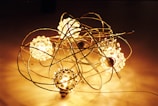 A decorative arrangement featuring several glowing spherical lights surrounded by intricate metallic wire structures. The spheres emit a warm, golden light, creating a cozy and artistic atmosphere. The wires are intertwined, forming a delicate and complex framework around the luminous orbs.