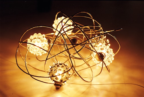 A decorative arrangement featuring several glowing spherical lights surrounded by intricate metallic wire structures. The spheres emit a warm, golden light, creating a cozy and artistic atmosphere. The wires are intertwined, forming a delicate and complex framework around the luminous orbs.