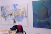 Two large abstract paintings are displayed on a white wall. The painting on the left features a blend of soft, pastel colors with sweeping brushstrokes that create dynamic, flowing forms. Various shades of blue, green, and hints of orange and yellow dominate the canvas. The painting on the right consists of deep blue and teal hues with subtle splashes of darker shades, conveying a sense of depth and movement. A chair with a pink garment draped over it, a coffee cup, papers, and an object that resembles a bag occupy the foreground on a light wooden table.