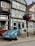 A vintage blue convertible car is parked on a cobblestone street in front of a traditional, half-timbered German house. The house features detailed trim and a sign above the windows. Hanging beside the car is a street lamp adorned with vibrant hanging flowers.