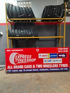 A display of tires is arranged on a yellow metal rack with two shelves. The upper and lower shelves each hold several rows of tires, neatly stacked and secured with white ties. Below the rack is a large red sign featuring the text 'EXPRESS TYRE SHOP' along with logos of various tire brands such as MRF, Michelin, Apollo Tyres, CEAT, and JK Tyre. The sign also includes contact information and the shop's location details.