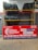 A display of tires is arranged on a yellow metal rack with two shelves. The upper and lower shelves each hold several rows of tires, neatly stacked and secured with white ties. Below the rack is a large red sign featuring the text 'EXPRESS TYRE SHOP' along with logos of various tire brands such as MRF, Michelin, Apollo Tyres, CEAT, and JK Tyre. The sign also includes contact information and the shop's location details.