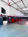 A spacious and organized automotive workshop featuring multiple hydraulic lifts arranged in a row. The walls are painted white with black and red accents. There is a red ceiling fan and a wall-mounted television. Various red tool cabinets and racks containing supplies are visible along the wall.