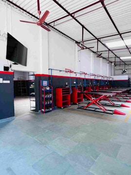 A spacious and organized automotive workshop featuring multiple hydraulic lifts arranged in a row. The walls are painted white with black and red accents. There is a red ceiling fan and a wall-mounted television. Various red tool cabinets and racks containing supplies are visible along the wall.
