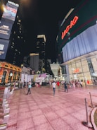 A bustling urban scene at night with a modern shopping complex featuring large illuminated signs. People are walking and socializing, some with shopping bags. The area is well-lit with neon lights and visible brand logos.