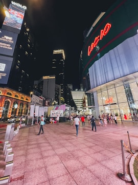 A bustling urban scene at night with a modern shopping complex featuring large illuminated signs. People are walking and socializing, some with shopping bags. The area is well-lit with neon lights and visible brand logos.
