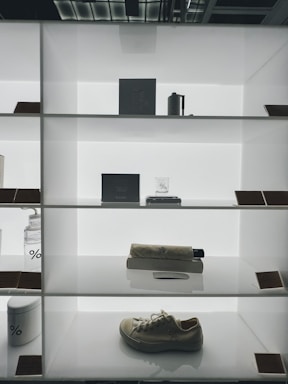 A minimalist display featuring several objects on a well-lit, white shelving unit. The shelves contain a mix of items including a white sneaker, a cylindrical container, a glass jar, a rolled fabric or umbrella, and a rectangular box. The background and shelving provide a sleek, modern look.