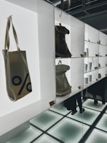 A modern display with shelves featuring various items, including canvas bags with a percentage sign, minimalist white cups, and metallic containers. The setting appears well-lit with bright, white backlighting and reflective surfaces creating a clean, contemporary look. There are a few people visible in the background, partially obscured by the shelves and reflections.