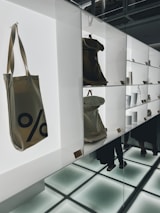 A modern display with shelves featuring various items, including canvas bags with a percentage sign, minimalist white cups, and metallic containers. The setting appears well-lit with bright, white backlighting and reflective surfaces creating a clean, contemporary look. There are a few people visible in the background, partially obscured by the shelves and reflections.