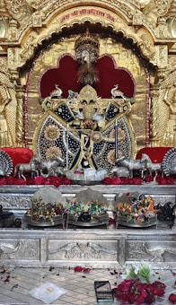 An ornately decorated altar features a central deity figure adorned with elaborate jewelry and vibrant colors. The entire setting is surrounded by intricate golden carvings, with red and black accents. Several small statues and offerings are placed on the lower platform, and a pattern of red roses and other flowers are spread in front.