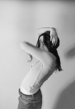 A woman is posing with her arms raised and bent, covering part of her face. She has long, dark hair and is wearing a light-colored top and denim pants. The background is a blank wall.