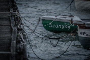 A small boat with the name 'Szunyog' and the number '63' is moored to a wooden dock with chains. The scene appears to be set in a coastal or lakeside area, with choppy waters and a slightly overcast atmosphere. The boat has a green hull and is positioned next to another partially visible boat.