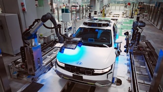 A modern automobile assembly line with a focus on a white car, which is surrounded by robotic arms. The setting is a highly automated factory environment, with various machinery and industrial equipment visible around. The car appears to be in the process of being assembled or inspected by the robotic systems.