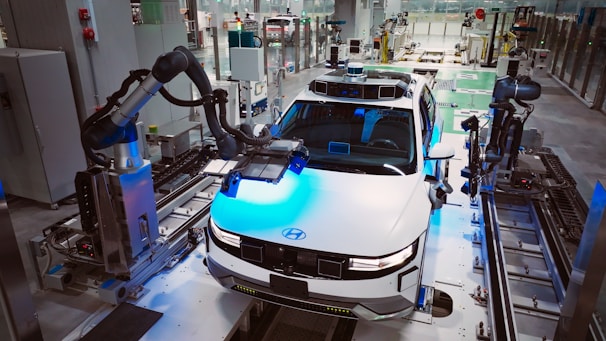 A modern automobile assembly line with a focus on a white car, which is surrounded by robotic arms. The setting is a highly automated factory environment, with various machinery and industrial equipment visible around. The car appears to be in the process of being assembled or inspected by the robotic systems.