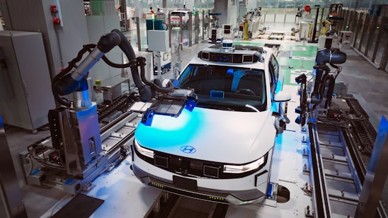 A modern automobile assembly line with a focus on a white car, which is surrounded by robotic arms. The setting is a highly automated factory environment, with various machinery and industrial equipment visible around. The car appears to be in the process of being assembled or inspected by the robotic systems.