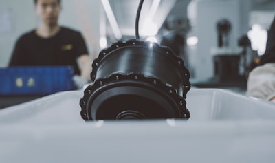 A large, cylindrical black machine part is placed under a bright light in a factory or workshop environment. In the background, a worker is slightly out of focus, indicating activity in the manufacturing process.