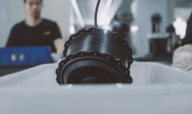 A large, cylindrical black machine part is placed under a bright light in a factory or workshop environment. In the background, a worker is slightly out of focus, indicating activity in the manufacturing process.