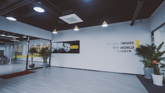A modern office lobby features a minimalist design with light wood flooring and white walls. Large glass doors are seen on the left, leading into another room. Several potted plants line the entryway, adding a touch of greenery. The ceiling has industrial-style exposed ductwork with pendant lights hanging down. A black and yellow sign with the words 'TRUCK RUN' is prominently displayed on one wall, while another wall has the phrase 'MOTOR DRIVES THE WORLD' in large letters.