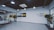 A modern office lobby features a minimalist design with light wood flooring and white walls. Large glass doors are seen on the left, leading into another room. Several potted plants line the entryway, adding a touch of greenery. The ceiling has industrial-style exposed ductwork with pendant lights hanging down. A black and yellow sign with the words 'TRUCK RUN' is prominently displayed on one wall, while another wall has the phrase 'MOTOR DRIVES THE WORLD' in large letters.