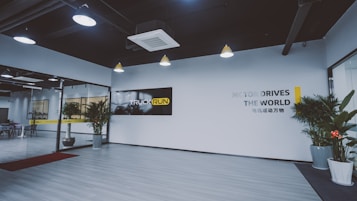 A modern office lobby features a minimalist design with light wood flooring and white walls. Large glass doors are seen on the left, leading into another room. Several potted plants line the entryway, adding a touch of greenery. The ceiling has industrial-style exposed ductwork with pendant lights hanging down. A black and yellow sign with the words 'TRUCK RUN' is prominently displayed on one wall, while another wall has the phrase 'MOTOR DRIVES THE WORLD' in large letters.
