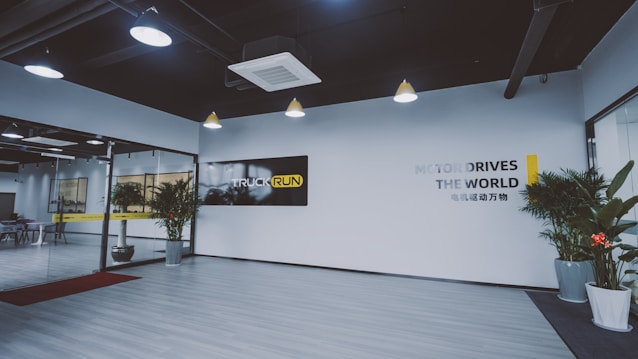 A modern office lobby features a minimalist design with light wood flooring and white walls. Large glass doors are seen on the left, leading into another room. Several potted plants line the entryway, adding a touch of greenery. The ceiling has industrial-style exposed ductwork with pendant lights hanging down. A black and yellow sign with the words 'TRUCK RUN' is prominently displayed on one wall, while another wall has the phrase 'MOTOR DRIVES THE WORLD' in large letters.